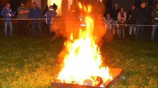 Im Vordergrund verbrennen die beiden Schlumpeln in ihrem Bett aus Metall, im Hintergrund sichert die Jugendfeuerwehr die Stelle ab, die Zuschauer machen Fotos von der Szene.