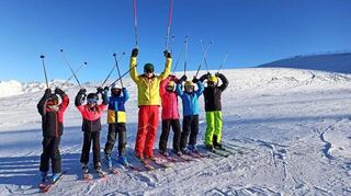 Die Kinder auf dem Bild freuen sich über die perfekten Fahrbedingungen mit klarem Himmel und weißen Pisten. In kleinen Gruppen üben sie das Slalomfahren und haben viel Spaß dabei.