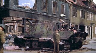 Diese Farbaufnahme zeigt das zerstörte deutsche Geschütz am ausgebrannten Haus Krebs. Ein GI der US-Army lässt sich von einem Kameraden fotografieren. Schwetzinger Jungs sind neugierig auf den Panzer geklettert.