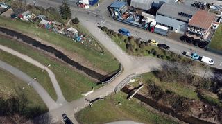 Die Blauseebrücke über den Kriegbach aus der Luft. Durch den oberen Teil des Bildes führt die Friedensstraße, von der aus die Zufahren zum Blausee abgehen. Wie aus der Luft gut zu erkennen ist, sind die Fuß- und Radwege von der Sanierung der Brücke nicht betroffen. Der auf dem Damm verlaufende Weg knickt vor der Brücke nach unten ab um anschließend auf dem Damm weiterzu-führen.