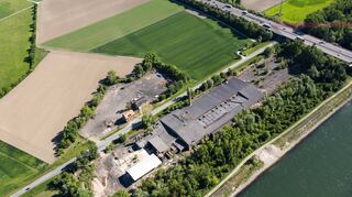 Das Luftbild zeigt, in welch besonderer Umgebung die ehemalige Ziegelei in der Gewann Herrenteich liegt: Nur wenige Meter entfernt fließt der Rhein vorbei, Hochwasserschutz gibt es nicht. Die Spannseile der Brücke der Autobahn 61 (r.) reichen bis ans Gelände heran. Im Dach der Halle sind dunkel die Löcher zu erkennen, durch die Regen eindringt. Rechts oben am Bildrand sind die Aussiedlerhöfe des Siegelhains sichtbar.