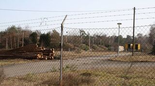 Eine trostlose Aussicht. Hinter dem Zaun, der mit Stacheldraht erhöht wurde, sieht man Baucontainer, Baumstämme und marode Asphaltwege.