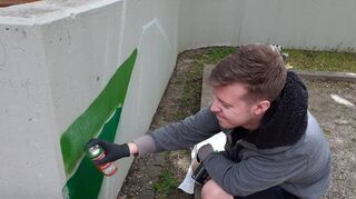 Finn Schuhmann beginnt das Kunstwerk an der Betonwand: Mit der grünen Farbe entsteht die Wiese – daneben sieht man schon leichte Umrisse für die Boulebahn.