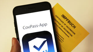 Das Logo der CovPass-App ist neben einem Impfbuch auf einem Smartphone zu sehen.