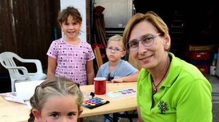 Maria Artner schminkt für Luisa (4) eine pinkfarbene Fee mit grünem Kleid. Noa und Jonas schauen interessiert zu.