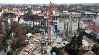 Gut abgeschirmte Baustelle: Von der Karlsruher Straße können Passanten nicht sehen, was sich hinter dem ersten Neubauabschnitt von St. Elisabeth (neben dem Baukran) tut. Der Anschluss des zweiten Abschnitts ist geschafft, der jetzt nach und nach in L-Form auf dem Gelände am Ufer des Kraichbachs (links) in die Höhe wachsen wird. Der Teil an der Stelle, wo der Kran steht, wird nicht unterkellert.
