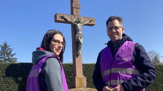 Die evangelische Pfarrerin Melanie Börnig und der katholische Diakon Heiko Wunder-ling stehen in ihren lilafarbenen Westen auf dem Brühler Friedhof für Gespräche mit Passanten bereit.