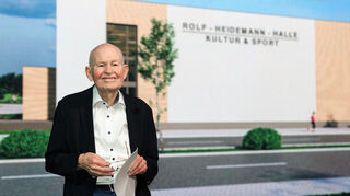 Rolf Heidemann vor der Leinwand, auf der der Name der neuen Kultur- und Sporthalle Neulußheim enthüllt wird. Sie wird seinen Namen tragen.