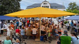 Wie ein Schiffssegel überspannt ein Sonnenschirm den Marktplatz: Darunter sitzen die Besucher des Gemeindefests der Evangelischen Kirchengemeinde.