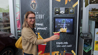 Lucia Orban hält vor dem Automaten eine Portion Pommes in den Händen. Auf einem Screen wird dem Kunden angezeigt, was er tun muss.