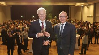 Bürgermeister Matthias Steffan (l.) und Oberbürgermeister Dr. René Pöltl stoßen symbolisch auf der Bühne im Lutherhaus an, im Hintergrund ist der Saal mit vielen Gästen zu sehen.