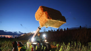 Landwirte zünden Mahnfeuer in Plankstadt. Ein weiterer Landwirt legt mit schwerem Gerät noch Strohballen ins Feuer.
