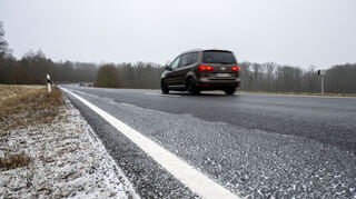 Ein Auto fährt über eine Straße, deren einer Rand mit Eis und leichtem Schnee bedeckt ist. Es herrscht Glatteisgefahr.