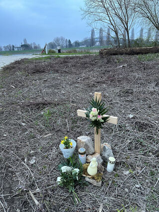Dieses Holzkreuz mit Kerzen und Blumen steht an der Stelle, an der die junge Frau gefunden wurde. Links die sogenannte Nato-Rampe und auf der anderen Rheinseite der Speyerer Müllberg.