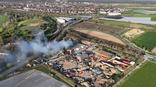 Der Rauch des Brands der Noba-Halle zieht auf die Bundesstraße 39, die von der Polizei vorsorglich gesperrt wird. Im Hintergrund das Gebäude der Firma Delvanis, wo es zuletzt im Jahr 2022 gebrannt hat. Zahlreiche Einsatzfahrzeuge der Feuerwehr sind auf dem Gelände zu erkennen.