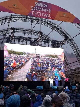 Mit Regenponchos ausstaffiert verfolgt das Publikum das Bühnengeschehen. SWR-Moderatorin Stephanie Haiber steht hier mit Richy Müller und Felix Klare auf der Bühne. "Sie sehen alle wie Zwerge in Ihren Ponchos aus", schickt Stephanie Haiber einen fröhlichen Gruß Richtung Publikum. Richy Müller witzelt, der Anblick erinnere an eine Versammlung vom Ku-Klux-Klan... Gelächter in den Gästereihen.