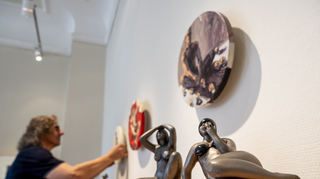 2405.24 Die Galerie Kulturraum Speyer zeigt im Zeitraum 26.05. - 21.07.24 die Ausstellung "Genau Betrachtet" mit Kunstwerken der Künstler Robert C. Rore und Eckhardt W. Rink. Hier von Rink vorne "Sitzende mit Zigarette" und dahinter "Ausblick". An der Wand hängen einige Tondi von Rore, von denen gerade Galerist Anton Bronich eins aufhängt