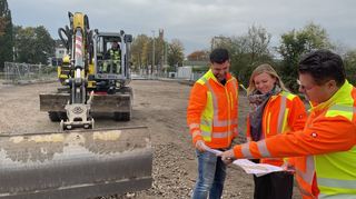 Luca di Candia (v. l.), Anna Krug und Kai Schemenauer schauen sich den Plan an, während die Bauarbeiter am Untergrund der Straße arbeiten.