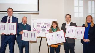 Ministerpräsident Alexander Schweitzer (l.) und die Repräsentanten der drei Städte präsentieren die neue SchUM-Briefmarke in der Mainzer Staatskanzlei.