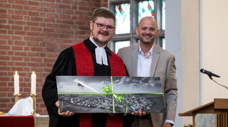 Eine Luftbildaufnahme von Oftersheim überreichte Bürgermeister Pascal Seidel (r) dem scheidenden Pfarrer Tobias Habicht mit den Worten: "Dies sei eine Drohnenaufnahme und die zeige den Ort aus einer Perspektive, wie ihn der liebe Gott sehen kann".