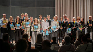 Lehrende und Lernende auf der Bühne; die Schülerinnen und Schüler erhalten jeweils eine Blume und ihr Studienbuch.